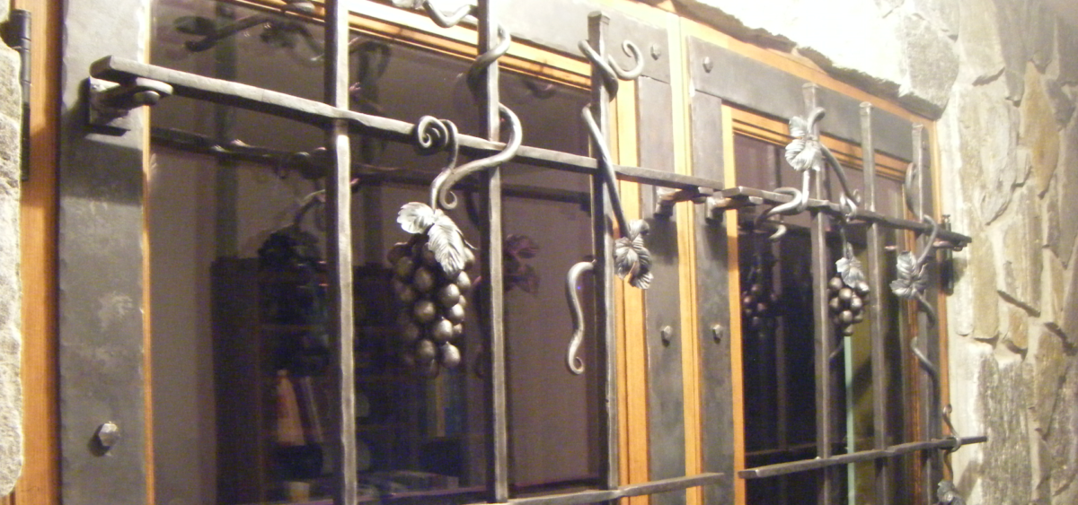 Double wine cellar doors with hammered bars. Vines wrapped around the bars and grapes hang.