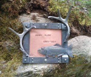 A custom made memorial plaque, with a steel frame and rivets. Around the frame is deer antlers and a fish made of steel. inside the frame is copper with stamped words.